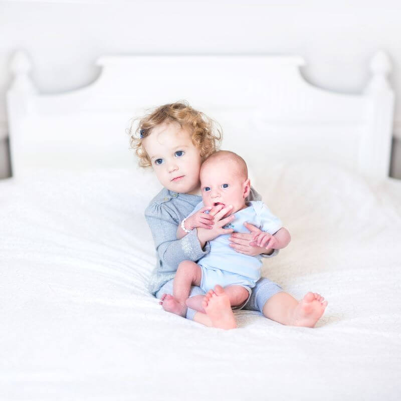 young child holding her baby sister while they both sit on a bed.