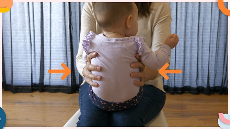 baby sitting on mothers lap, with her hands gently holding the baby by her sides. Baby is sitting upright, with her head straight