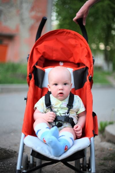 Baby boy sitting in a red stroller wearing a five point harness, facing the camera.