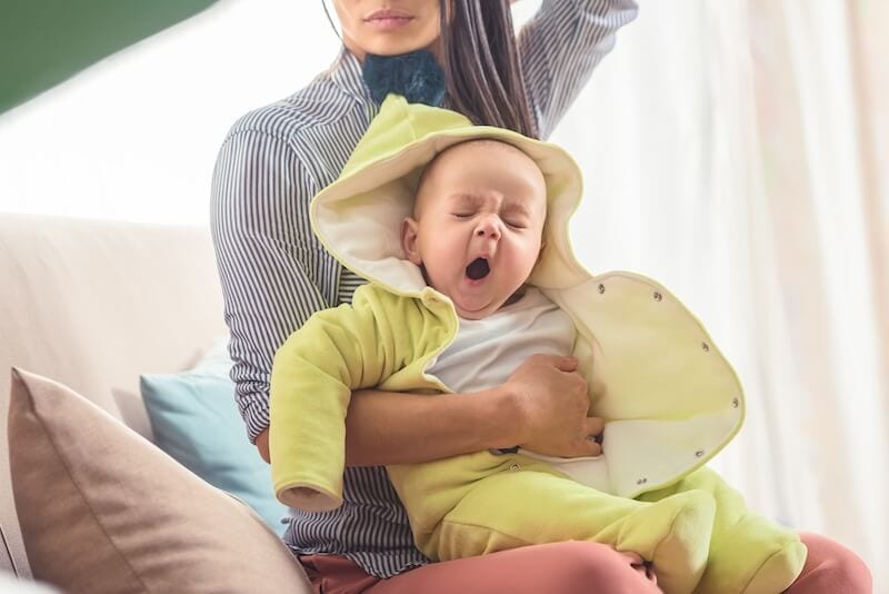 baby wearing a yellow hooded jacket yawning in his mothers arms due to being overtired