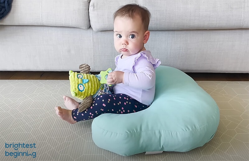 baby girl wearing a pink shirt and holding a soft owl toy is sitting in a green boppy pillow looking at the camera