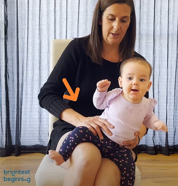 woman sitting on a white chair with a baby girl wearing a pink shirt sitting on her lap facing forwards. Woman gently presses on baby's right hip to help bring her back up to an upright position.