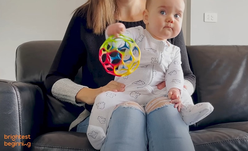 baby in white onesie sitting on mothers lap facing outwards. Baby holding a round toy ball. Mother holding baby down around hips for support