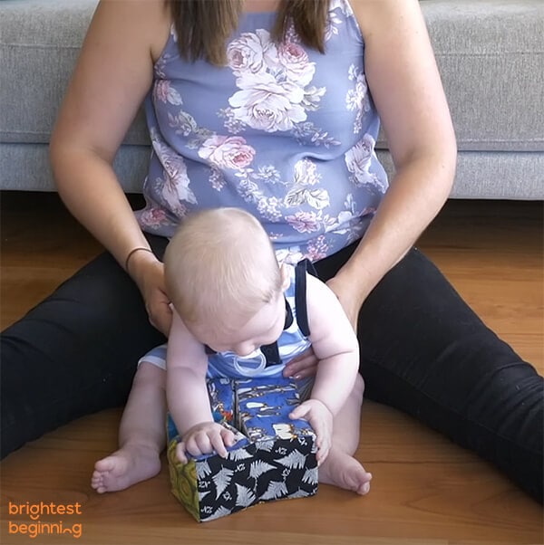 infant sits between a woman's legs facing forwards with his hands resting on a soft, square toy for support